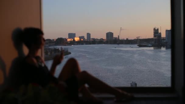 Девушка сидит у окна на фоне города и моря — стоковое видео