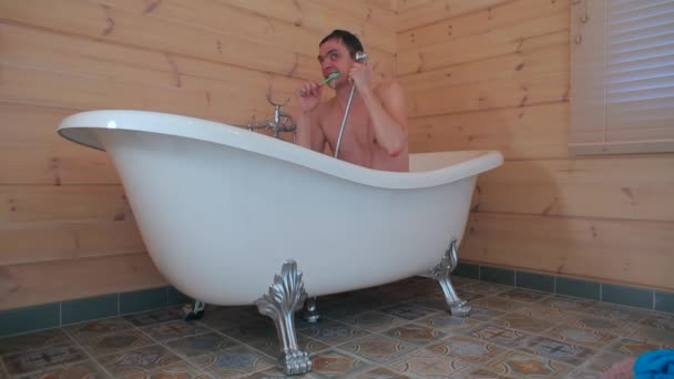 Парень, сидящий в ванной, чистит зубы и говорит по телефону дешево . — стоковое видео