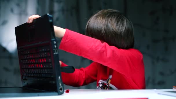 Un niño sentado en una mesa examina la parte posterior de un portátil . — Vídeo de stock