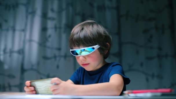 Ένα αγόρι με εικονικά γυαλιά που κάθεται σε ένα τραπέζι παίζει ένα βιντεοπαιχνίδι σε ένα smartphone. — Αρχείο Βίντεο