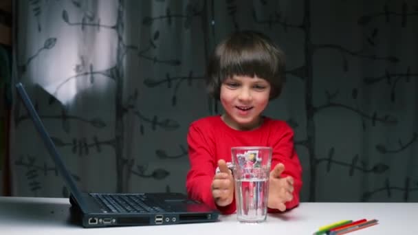 En pojke som sitter vid ett bord häller vatten från ett glas på en laptop. — Stockvideo