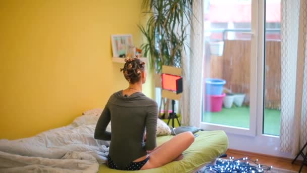Uma mulher senta-se em uma cama em um quarto, medita enquanto olha pela janela . — Vídeo de Stock