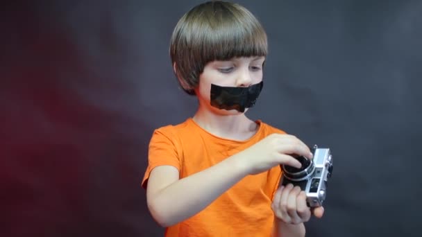 En pojke med svart tejp över munnen undersöker en gammal kamera. — Stockvideo