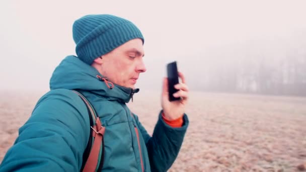 Um homem de chapéu está falando em um celular enquanto está em um campo nebuloso . — Vídeo de Stock