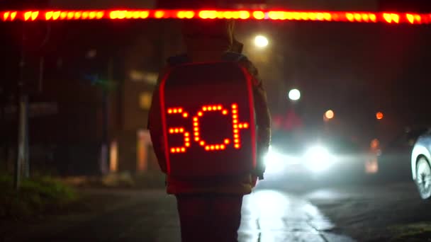 Een schooljongen met een lichtgevende rugzak staat 's avonds op de oversteekplaats op school. — Stockvideo