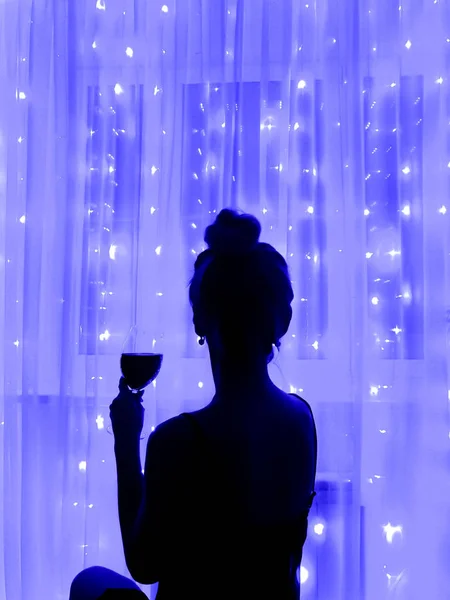 Silhouette of a girl with a glass of wine on the background of luminous lights of a garland.