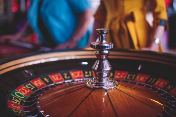 A close-up vibrant image of multicolored casino table with roulette in motion, with the hand of croupier, and a group of gambling rich wealthy people 