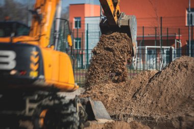 Sarı ağır kazıcı kumu kazıyor ve yol çalışmaları sırasında çalışıyor, yeni yolun inşaatı sırasında kumu boşaltıyor ve etrafta işçiler var.