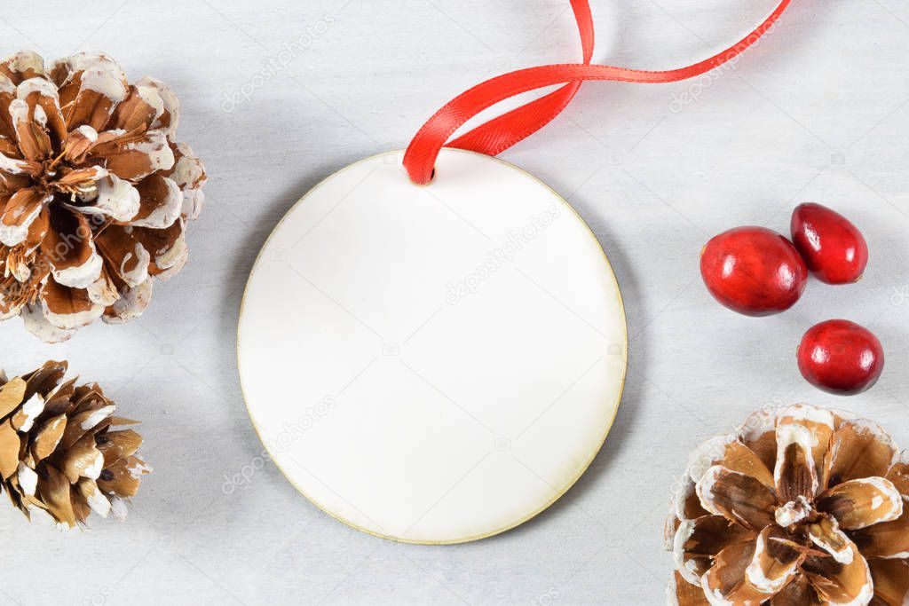 Christmas Ornament Mock Up featuring a round Christmas ornament tied with a red ribbon atop a light gray wood background. Pine cones and fresh cranberries surround the round ornament.