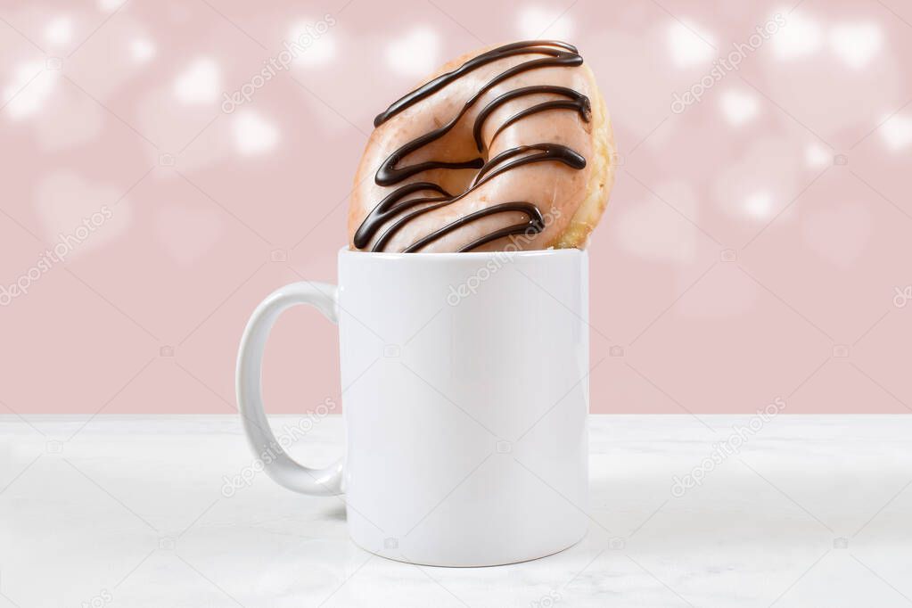 11 ounce white coffee mug filled with a glazed chocolate ice donut. Bokeh hearts glow in the pink background.