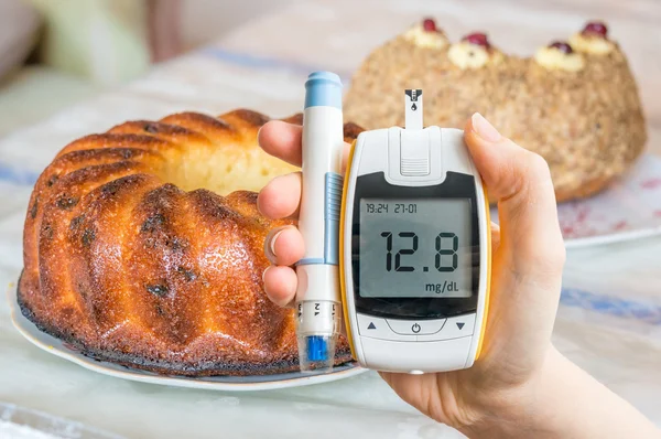 Diabetes, diet and unhealthy eating concept. Hand holds glucometer and cakes and sweets in background.