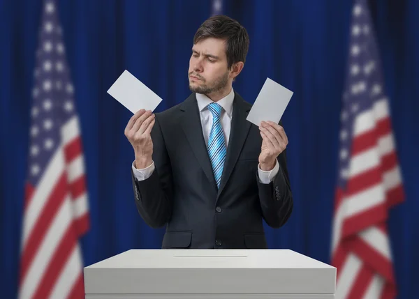 Election in United States of America. Undecided voter holds envelopes in hands above vote ballot and making decision. USA flags in background. Democracy concept. — Stock Photo, Image