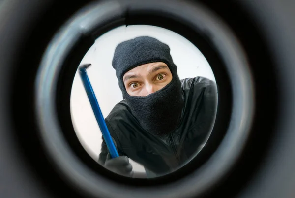Zobrazit prostřednictvím pipehole. Zloděj nebo lupič maskovaný s balaclava drží páčidlo za dveřmi. — Stock fotografie