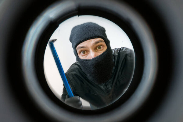 View through pipehole. Thief or burglar masked with balaclava is holding crowbar behind the door.