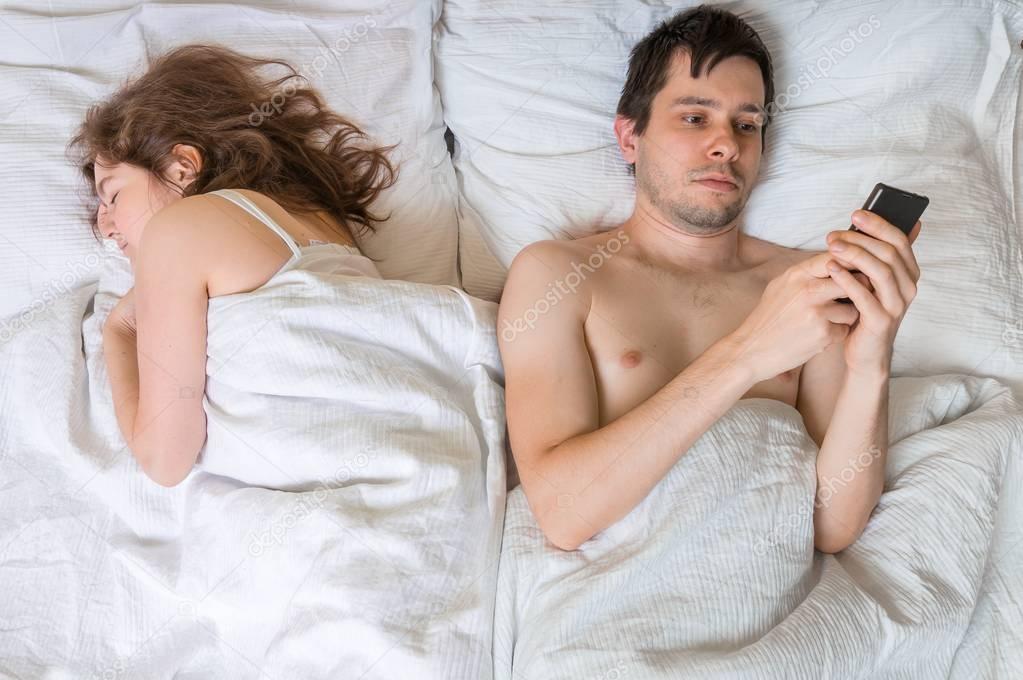 Young man is texting with someone using phone while his wife is sleeping near him. Cheating concept.