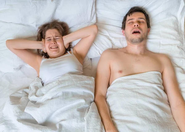 La joven está acostada con su marido en la cama. El hombre ronca demasiado fuerte. La mujer se cubre los oídos . — Foto de Stock
