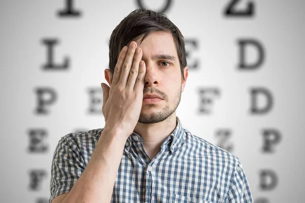 O jovem está a cobrir o rosto com a mão e a verificar a visão. Gráfico para testes de visão ocular em segundo plano . — Fotografia de Stock