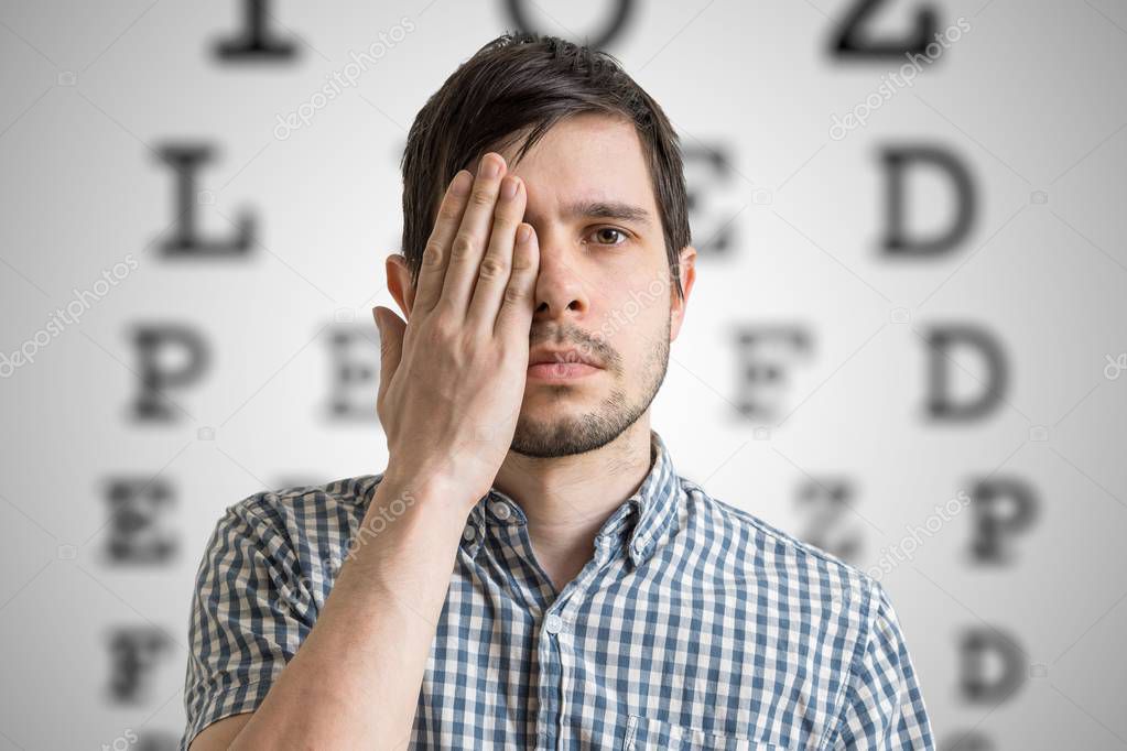 Young man is covering his face with hand and checking his vision. Chart for eye sight testing in background.