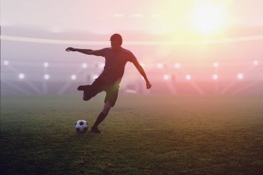 Futbolcu günbatımında stadyumda bir topa vuruyor.