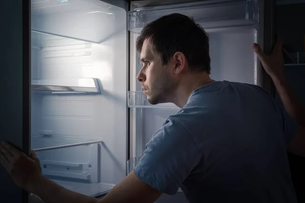 Homme affamé est à la recherche de nourriture dans le réfrigérateur vide la nuit . — Photo