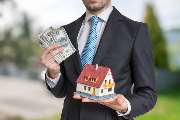 Agente inmobiliario está sosteniendo pequeña casa y dinero en las manos . — Foto de Stock