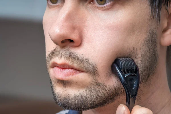 Facial hair care concept. Young man is using derma roller  on be — Stock Photo, Image