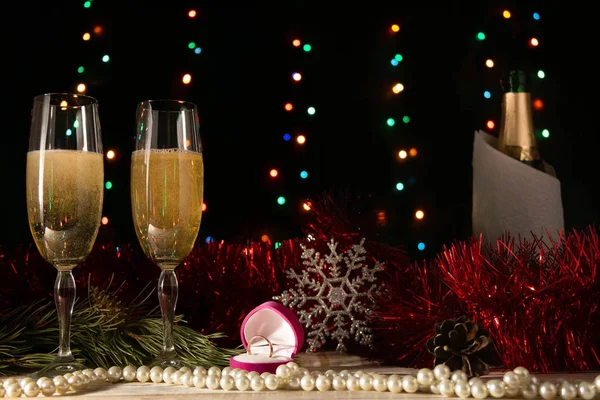 Romantic Christmas and New Year dinner of a couple in love with two glasses of champagne and a wedding ring. Red tinsel and spruce branch on a table on a dark background with blurred lights.