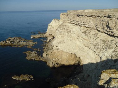 Yüksek engebeli bir kıyı kayalığı ve zar zor fark edilen bir tekne keskin kayaların arasında duruyor. Güneşli bir gün ve denize yolculuk.