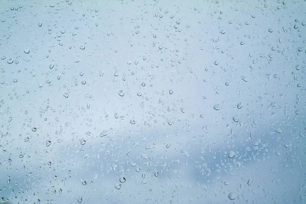 Close-up raindrops on the glass and the sky outside the window. Blue textured abstract background with water drops and rainy weather. Transparent wet non-uniform surface, condensation effect.