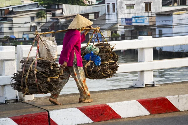 Barefoot Vietnamese mature woman in conical Asian hat carrying wood in busy street near Chinese quarter on February 13, 2012 in My Tho, Vietnam. — Stock Photo, Image