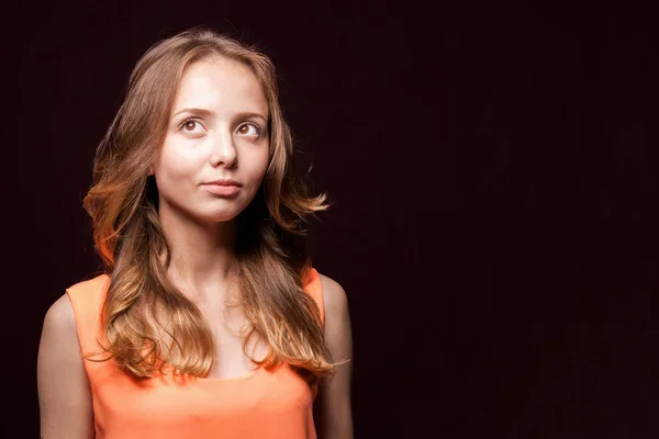 Dromerig leuk meisje in een oranje blouse op een zwarte achtergrond. — Stockfoto