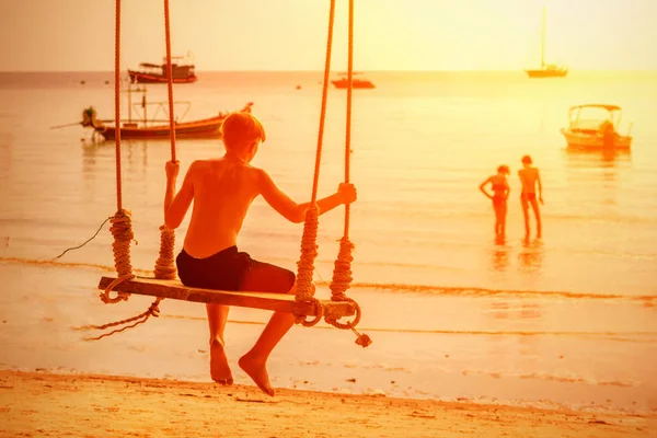 Le garçon oscille sur la balançoire de la plage au coucher du soleil. Vue de l'arrière . — Photo