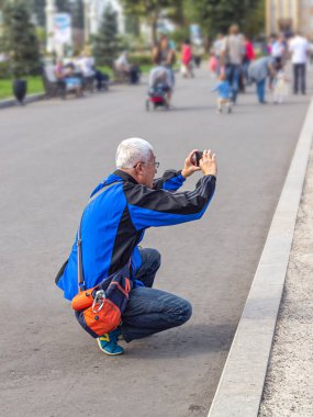 Gri saçlı, yaşlı bir Asyalı adam yere çömelmiş telefonuyla fotoğraf çekiyor.