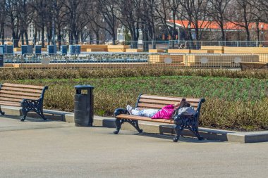 Kot pantolonlu, spor ayakkabılı ve pembe ceketli kız başının altında sırt çantasıyla bankta uzanıyor ve akıllı telefona bakıyor. Ilık bahar sabahı