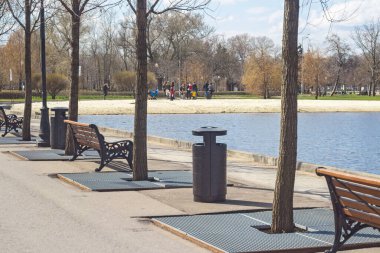 Göl kenarındaki ara sokakta ağaçlar, ahşap koltuklu dökme demir banklar ve siyah metal vazolar var. Kumsalda çocuklu bir grup ebeveyn