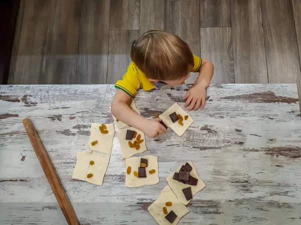 a small child standing on a stand helps in the kitchen at home, rolls puff pastry for buns / pies and puts in them a filling of pieces of chocolate and raisins.Design of cards, screensavers, covers