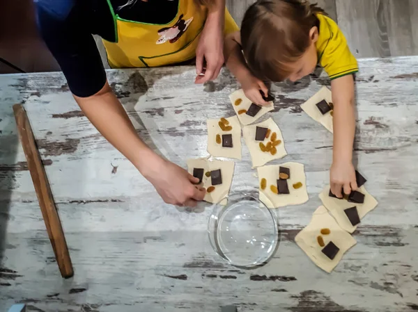 a small child standing on a stand helps in the kitchen at home, rolls puff pastry for buns / pies and puts in them a filling of pieces of chocolate and raisins.Design of cards, screensavers, covers