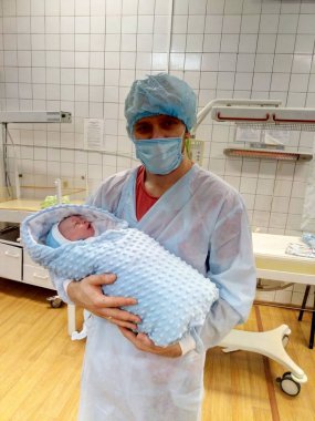 Tek kullanımlık tıbbi giysili açık tenli bir adam yeni doğmuş bir bebeği doğumhanede tutuyor..