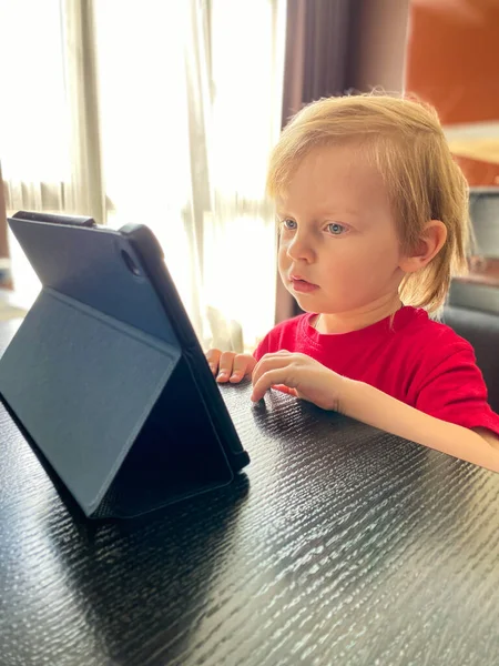 A fair-skinned little child sitting at a table in the kitchen, self-learning using a tablet.