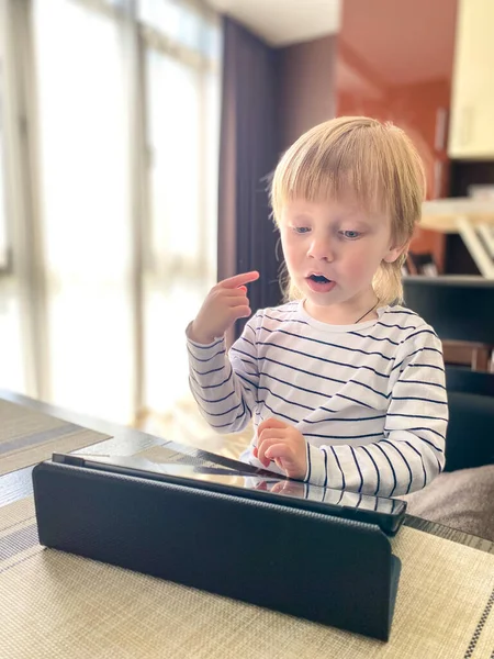 A fair-skinned little child sitting at a table in the kitchen, self-learning using a tablet.Defocus light background.