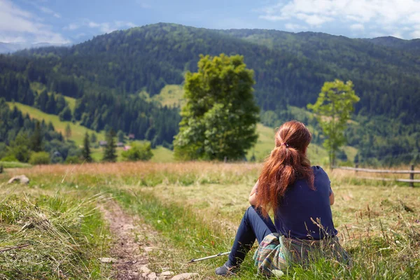 Redhead woman hiker in a dark blue t-shirt sitting on a meadow, admiring a mountain view looking out over distant ranges and fir-tree forest. — Stock Photo, Image