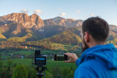 Arkaya bakan genç adam telefonda fotoğraf çekiyor. Dağ sıralarında günbatımını kaydediyor.