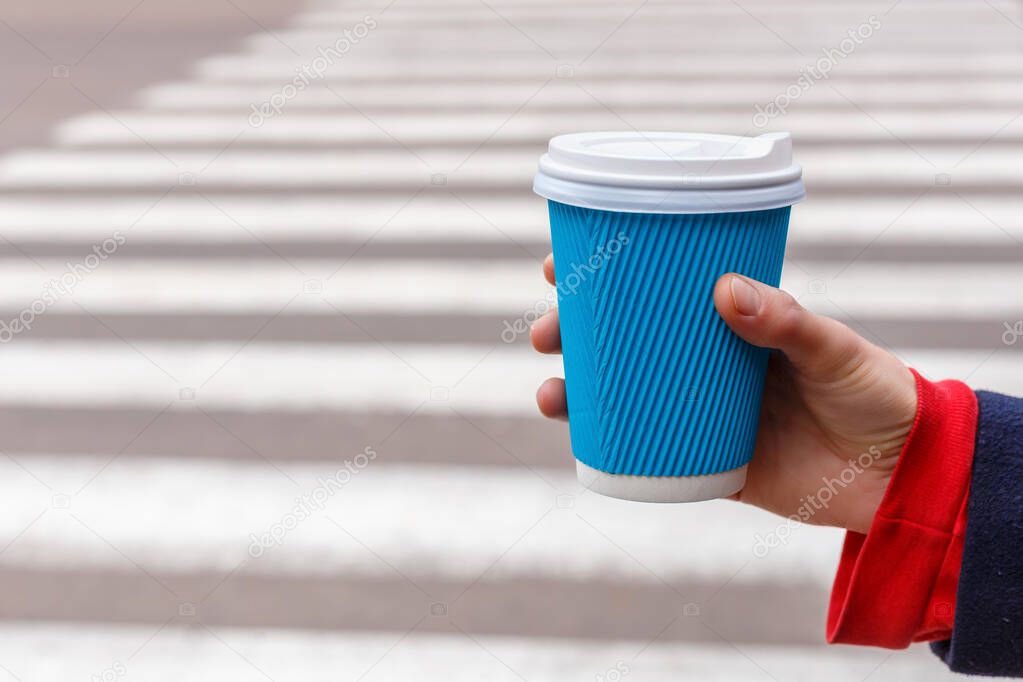 Close-up woman holding a blue paper cup of coffee in hand on zebra crossing background.