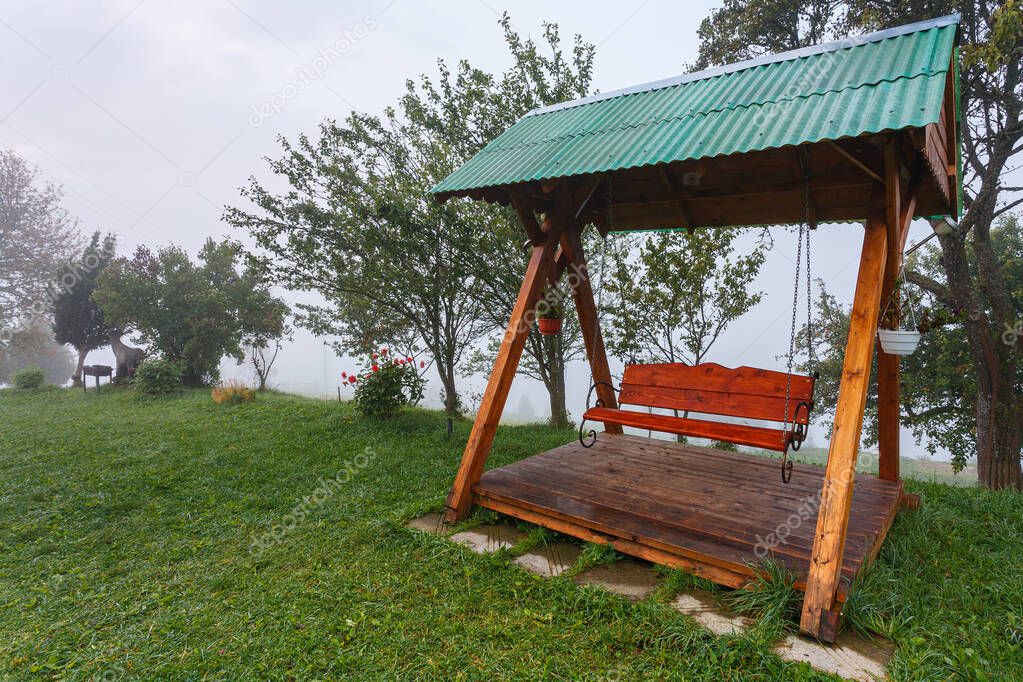 Red swing bench with green roof in forest at foggy autumn morning with trees and vivid green grass