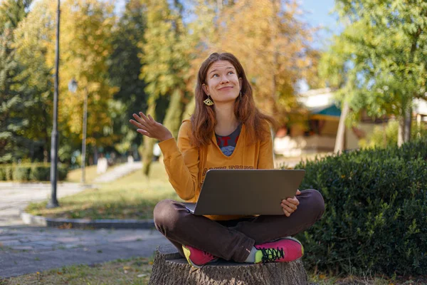 Νεαρό κορίτσι με κίτρινη κουκούλα με φορητό υπολογιστή, κοιτάζει ψηλά, εκφράζει συναισθήματα έκπληξη και αναρωτιέται σε εξωτερικούς χώρους, κάθεται στο κούτσουρο στο πάρκο φθινόπωρο Εικόνα Αρχείου