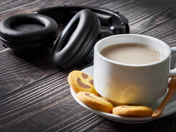 Μεγάλο λευκό φλιτζάνι καφέ, μαύρα ακουστικά και κίτρινα μπισκότα με χαρούμενα χαμόγελα Εικόνα Αρχείου
