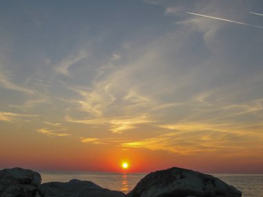 Deniz kıyısında şafak vakti, ön planda taşlarla, mavi gökyüzü ve