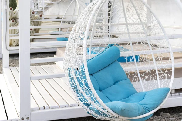 Modern design white luxury hanging rattan wicker chair egg form with cozy blue pillows inside on the beach