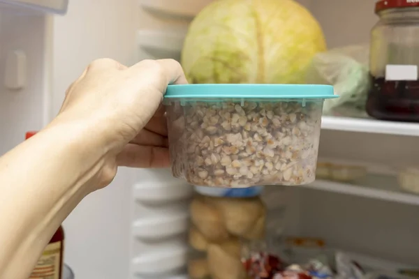 Uma mão de uma mulher de dieta tomando um recipiente de plástico com trigo mourisco cozido de um refrigerador, congelador, perdendo conceito de peso — Fotografia de Stock