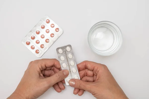 Woman taking pills, female hands holding a blister of white pills, capsules and a glass of water on white table, healthcare and flu treatment concept